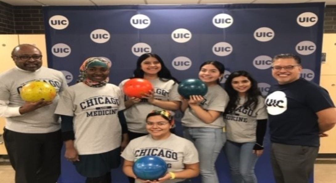 a diverse group of people holding bowling balls, posing in front of a backdrop with UIC logos